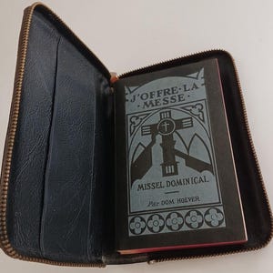 May include: A black leather zipped case containing a small book. The book cover is black with a blue and white design and the text "J'OFFRE LA MESSE. MISSEL DOMINICAL. Par Dom HOEVER".