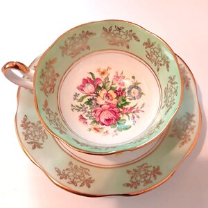 May include: A vintage teacup and saucer set. The cup and saucer are a pale green with gold trim and floral patterns. The interior of the cup features a bouquet of pink, red, and yellow flowers. The handle of the cup is gold.
