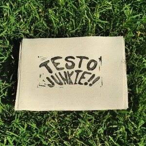 Könnte beinhalten: Ein rechteckiger, cremefarbener Stoff mit den Worten "TESTO JUNKIE!!" in schwarzen Blockbuchstaben. Der Stoff liegt vor einem Hintergrund aus grünem Gras.