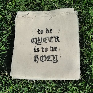 Könnte beinhalten: Ein quadratischer, cremefarbener Stoff mit dem Text "to be QUEER is to be HOLY" in schwarzer gotischer Schrift. Der Stoff hat rohe Kanten und liegt auf grünem Gras.