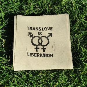 May include: A square, off-white fabric patch with the words "TRANS LOVE IS LIBERATION" printed in black. The design includes intertwined gender symbols. The patch is set against a background of green grass.