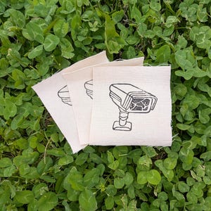 Puede incluir: Tres cuadrados de tela blanquecina con un dibujo lineal negro de una cámara de seguridad. Los cuadrados están colocados sobre un lecho de tréboles verdes. La cámara se representa con una lente detallada y una base robusta.