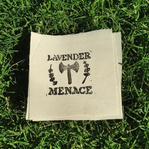 Può includere: Toppa quadrata di tessuto bianco sporco con le parole "LAVENDER MENACE" stampate in nero, insieme a un'ascia e illustrazioni floreali. La toppa è appoggiata su un letto di erba verde.