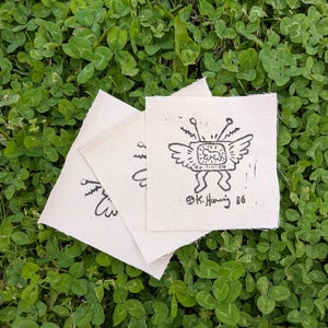 May include: Three small, off-white fabric squares with a black ink drawing of a television with wings, antennae, and legs. The text "K. Haring 86" is printed on the bottom right corner. The squares are on a bed of green clover.