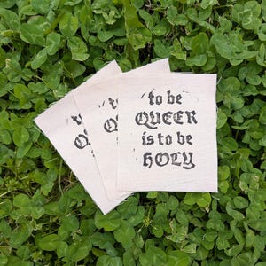 May include: Three off-white fabric patches with the text "to be QUEER is to be HOLY" in black gothic lettering. The patches are arranged on a bed of green clover.