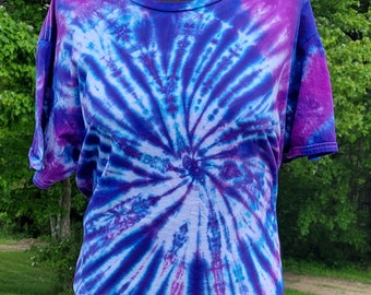 Op de afbeelding: Een tie-dye T-shirt met een draaiende print in tinten paars, blauw en wit.