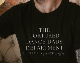 May include: Black t-shirt with the text "THE TORTURED DANCE DADS DEPARTMENT" and the phrase "ALL'S FAIR IN love AND competitions" in a white, elegant font. The person is holding a bottle of beer.