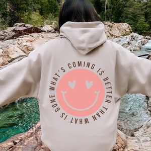 Puede incluir: Una sudadera con capucha beige con un gráfico de una cara sonriente rosa en la espalda. El gráfico dice "What's gone is what's coming is better than".