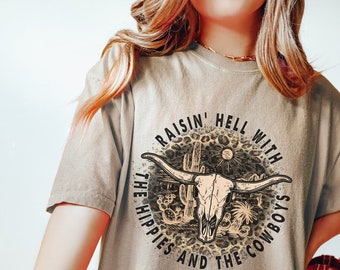 Könnte beinhalten: Ein hellbraunes T-Shirt mit einem Grafikdruck eines Schädels mit Hörnern, Kakteen und dem Text "Raisin' Hell With The Hippies And The Cowboys".