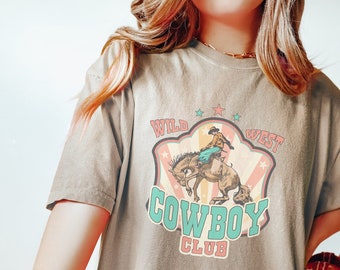 Könnte beinhalten: Ein hellbraunes T-Shirt mit einer Grafik im Vintage-Stil, die einen Cowboy auf einem buckelnden Pferd zeigt. Die Grafik ist von einem roten, weißen und blauen Sternenausbruch umgeben und trägt den Text "Wild West Cowboy Club".
