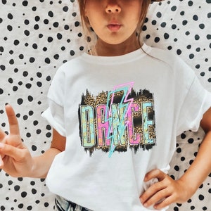 May include: White t-shirt with a graphic design of the word "DANCE" in colorful letters with a lightning bolt behind it. The graphic is on a brown and black leopard print background.