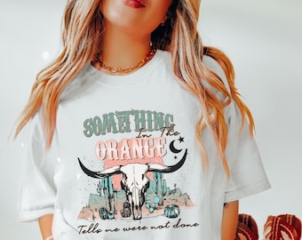 Könnte beinhalten: Weißes T-Shirt mit einem Grafikdruck eines Kuhschädels, Kakteen und dem Text "Something in the Orange Tells me we're not done".