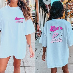 Op de afbeelding: Wit oversized T-shirt met roze retro-lettertype tekst aan de voorkant die "Dance Mom" zegt en aan de achterkant die "Let's Go Girls" zegt met roze sterren.