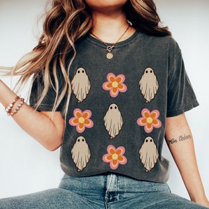 May include: A dark gray t-shirt with a pattern of white ghosts and pink and orange flowers. The shirt is being worn by a person sitting down.