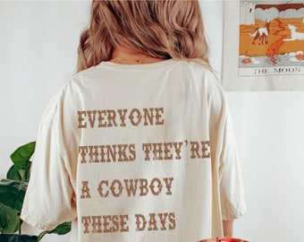 Könnte beinhalten: Ein beiges T-Shirt mit dem Text "Everyone thinks they're a cowboy these days" auf der Rückseite. Die Vorderseite des Shirts zeigt einen kleinen Schädel, der einen Cowboyhut trägt.