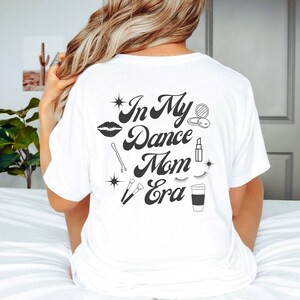 May include: White t-shirt with the text "In My Dance Mom Era" in black script. The shirt features various black and white illustrations of makeup and beauty items, including a lipstick, eyelashes, a makeup brush, and a hair clip.