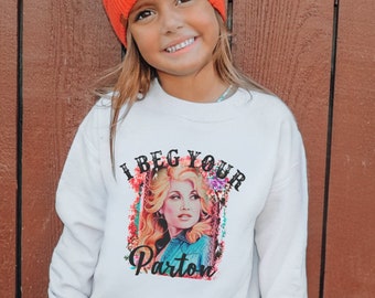 May include: A young person wearing an orange beanie and a white sweatshirt. The sweatshirt features a colorful graphic of a woman with the text "I Beg Your Parton". The background is a brown wooden wall.