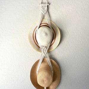 May include: A white macrame hat rack with two straw hats hanging from it. The rack has a long tassel hanging from the bottom.