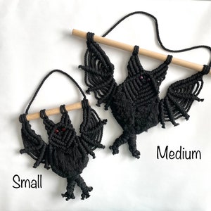 May include: Two black macrame bat wall hangings suspended from light-colored wooden dowels with attached hanging cords. The smaller bat features red bead eyes, while the larger medium bat has dark purple bead eyes. The text Small and Medium is visible.