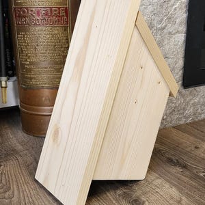 K&ouml;nnte beinhalten: Ein kleines, unfertiges Vogelhaus aus Holz mit einem Satteldach. Das Vogelhaus besteht aus hellfarbenem Holz und hat ein einfaches, rechteckiges Design. Das Vogelhaus steht auf einer Holzoberfl&auml;che.