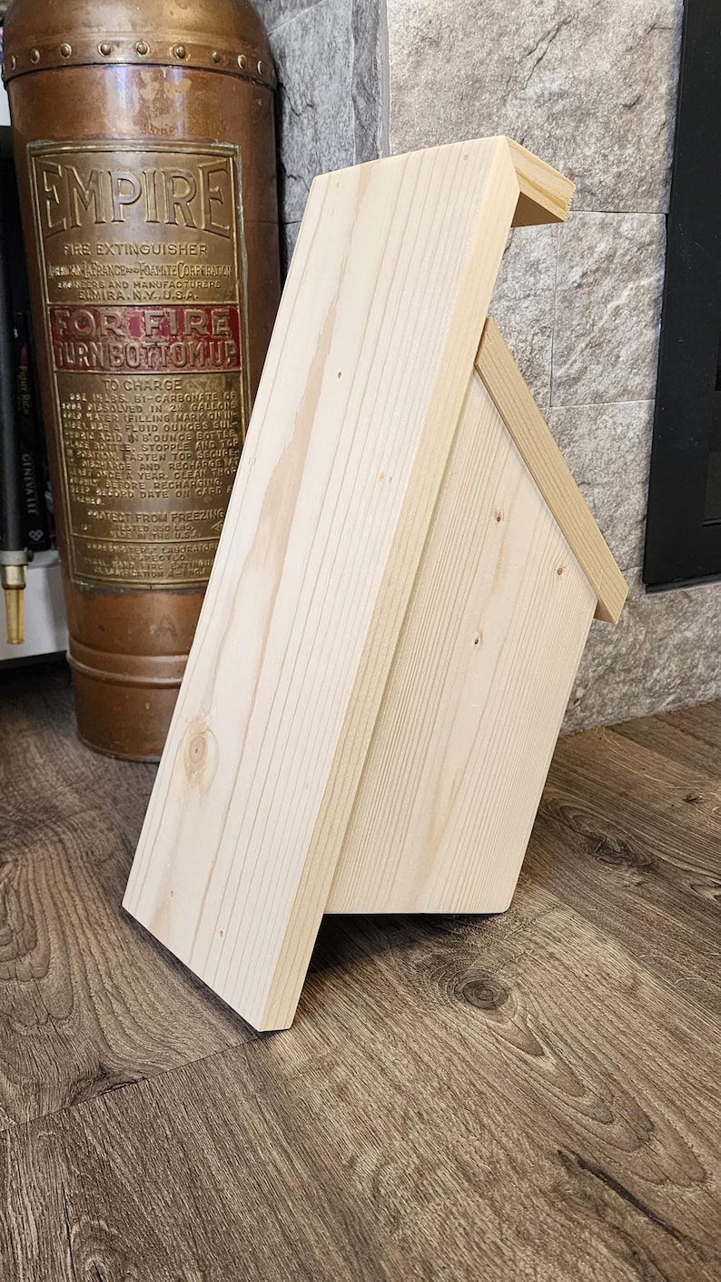 K&ouml;nnte beinhalten: Ein kleines, unfertiges Vogelhaus aus Holz mit einem Satteldach. Das Vogelhaus besteht aus hellfarbenem Holz und hat ein einfaches, rechteckiges Design. Das Vogelhaus steht auf einer Holzoberfl&auml;che.