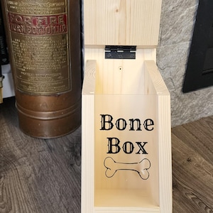 K&ouml;nnte beinhalten: Helle Holzhundekeksdose mit Klappdeckel. Auf der Vorderseite der Box steht "Bone Box" und eine Knochengrafik. Der Deckel ist mit "Uncle Bone's" und einem dekorativen Emblem graviert. Die Box ist aus Holz.