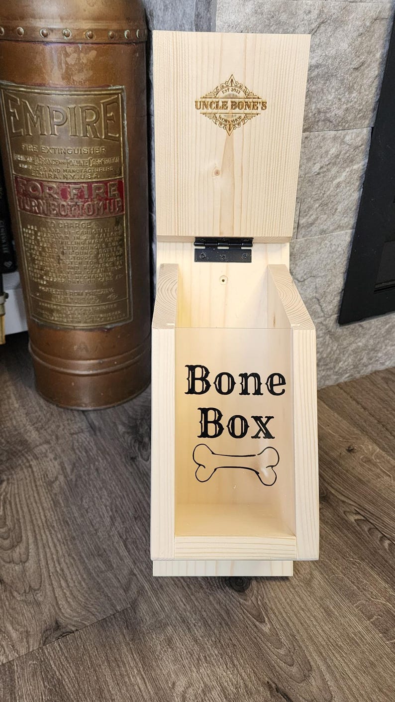K&ouml;nnte beinhalten: Helle Holzhundekeksdose mit Klappdeckel. Auf der Vorderseite der Box steht "Bone Box" und eine Knochengrafik. Der Deckel ist mit "Uncle Bone's" und einem dekorativen Emblem graviert. Die Box ist aus Holz.
