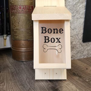 K&ouml;nnte beinhalten: Eine Hundekeksdose aus Holz mit einer transparenten Frontplatte. Die Worte "Bone Box" und eine Knochengrafik sind auf die Platte gedruckt. Die Box ist aus hellem Holz gefertigt und hat ein kleines Dach. Ein antiker Feuerl&ouml;scher ist im Hintergrund.