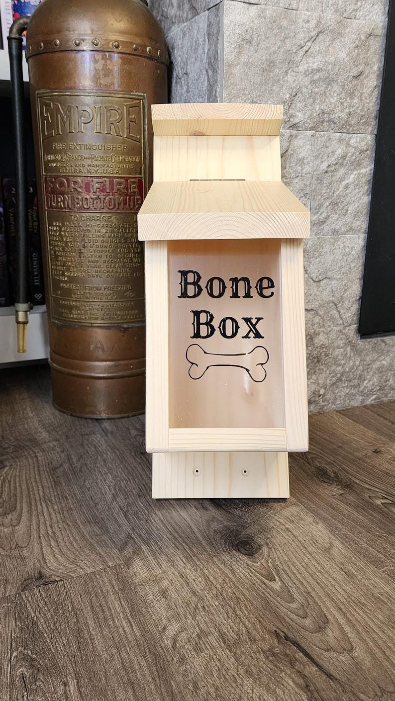 K&ouml;nnte beinhalten: Eine Hundekeksdose aus Holz mit einer transparenten Frontplatte. Die Worte "Bone Box" und eine Knochengrafik sind auf die Platte gedruckt. Die Box ist aus hellem Holz gefertigt und hat ein kleines Dach. Ein antiker Feuerl&ouml;scher ist im Hintergrund.