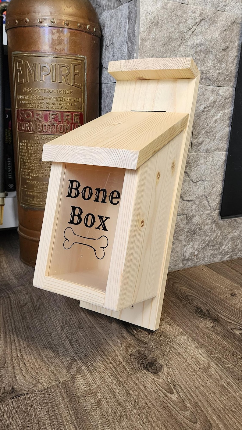 K&ouml;nnte beinhalten: Eine Hundeknochenbox aus Holz mit einer transparenten Frontplatte. Die Worte "Bone Box" und eine Knochengrafik sind auf der Vorderseite aufgedruckt. Die Box ist aus hellem Holz gefertigt und hat ein schr&auml;ges Dach.