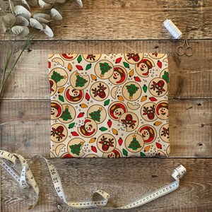 Puede incluir: Un papel de regalo de color crema con un patr&oacute;n repetitivo de galletas de Navidad. Las galletas est&aacute;n decoradas con &aacute;rboles verdes, luces rojas y caras de mu&ntilde;eco de nieve y reno.