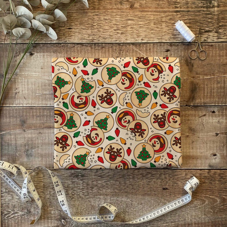 Puede incluir: Un papel de regalo de color crema con un patr&oacute;n repetitivo de galletas de Navidad. Las galletas est&aacute;n decoradas con &aacute;rboles verdes, luces rojas y caras de mu&ntilde;eco de nieve y reno.
