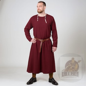 May include: A burgundy-colored medieval tunic with long sleeves and a round neckline. The tunic is cinched at the waist with a brown leather belt. The model is wearing brown socks and black leather shoes. The tunic is suitable for historical reenactments or themed events.