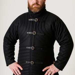 May include: A black quilted coat with a high collar and leather straps with metal buckles. The coat is buttoned up the front and has a long length. The person is wearing the coat and has their hands on their hips.