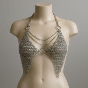 Könnte beinhalten: Silbernes Kettenhemd-Haltertop mit dreieckigem Design. Das Top besteht aus kleinen Metallringen, die miteinander verbunden sind und ein netzartiges Gewebe bilden. Die Halterriemen und dekorativen Ketten sind mit Metallringen befestigt.