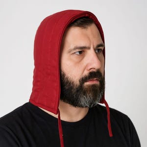 May include: A red padded hood with a drawstring closure. The hood is designed with vertical stitching and is made of a solid, dark red fabric. The hood is worn over a black shirt.