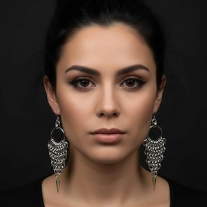 May include: Silver chainmail earrings with a circular top and a fringed design. Each earring has a gold-coloured, pointed bottom. The earrings are worn by a person with dark hair and a black top against a dark background.