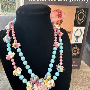 Puede incluir: Un collar de doble cadena con cuentas de color azul pastel y rosa y un gran colgante floral. El colgante está hecho de flores rosas, blancas y amarillas con un gran centro rosa. El collar se muestra en un soporte de terciopelo negro. La imagen está acompañada de un libro titulado "Miriam Haskell Jewelry".