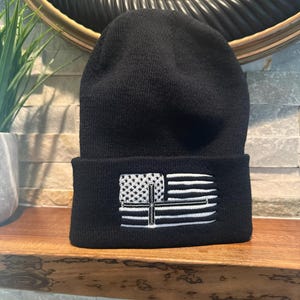 May include: A black beanie with a white embroidered design of a flag and a cross. The beanie is made of a soft, knitted material and has a folded brim. The design features stars and stripes.