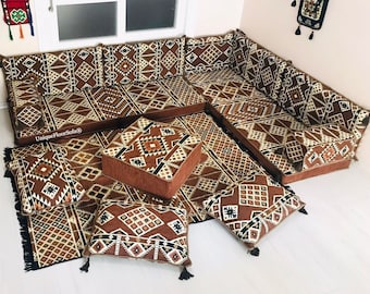 May include: A brown and beige patterned floor cushion seating set with a geometric design. The set includes a large L-shaped sofa, a square ottoman, and several smaller square cushions. The cushions are all covered in a soft, plush fabric and have tassels on the corners.