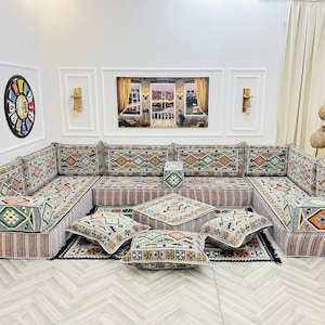 May include: A gray and white sectional sofa with colorful patterned cushions. The sofa is arranged in a U-shape and has a variety of throw pillows in different patterns and colors. The sofa is set against a white wall with a window behind it.