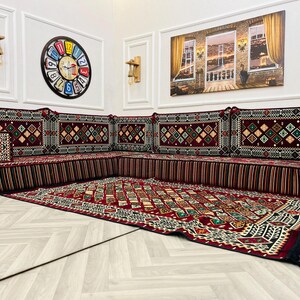 May include: A traditional red and black patterned diwan seating set with a matching rug. The diwan is made up of multiple cushions with intricate embroidery and fringe details. The rug has a geometric design with a black border.