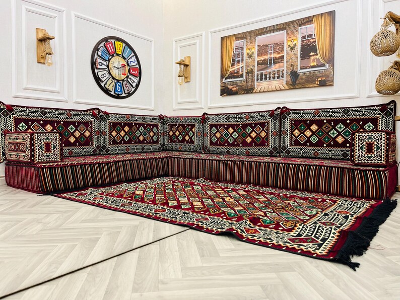 May include: A traditional red and black patterned diwan seating set with a matching rug. The diwan is made up of multiple cushions with intricate embroidery and fringe details. The rug has a geometric design with a black border.