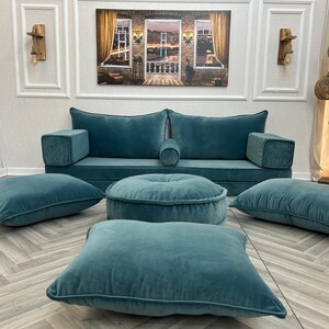 May include: A teal blue sofa with four matching teal blue cushions and a round teal blue ottoman. The sofa is against a white wall with a picture hanging above it.