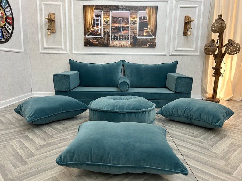 May include: A teal blue sofa with four matching teal blue cushions and a round teal blue ottoman. The sofa is against a white wall with a picture hanging above it.