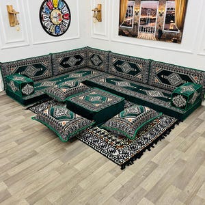 Puede incluir: Un sofá seccional verde con cojines estampados y una mesa de centro a juego. El sofá está dispuesto sobre una alfombra estampada, creando una zona de estar acogedora. Obras de arte murales y lámparas decorativas completan la decoración de la habitación.