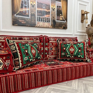 May include: A red and white striped floor cushion with matching pillows. The pillows have a green, red, and white geometric pattern. The set includes a small square cushion with tassels. The background features a painting of a cityscape.