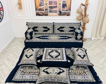 May include: A black and gold patterned divan with matching throw pillows and floor cushions. The divan is made of fabric and has a traditional design.