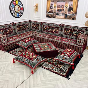 May include: A traditional red and gold patterned divan with matching cushions and throw pillows. The divan is set on a patterned rug with a decorative clock on the wall behind it.