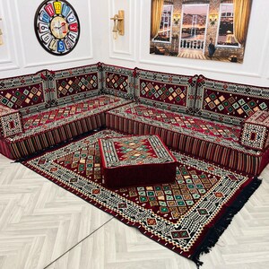 May include: A red and gold patterned divan with a matching patterned rug and a red and gold patterned ottoman. The divan is made up of multiple cushions and has a fringe border.